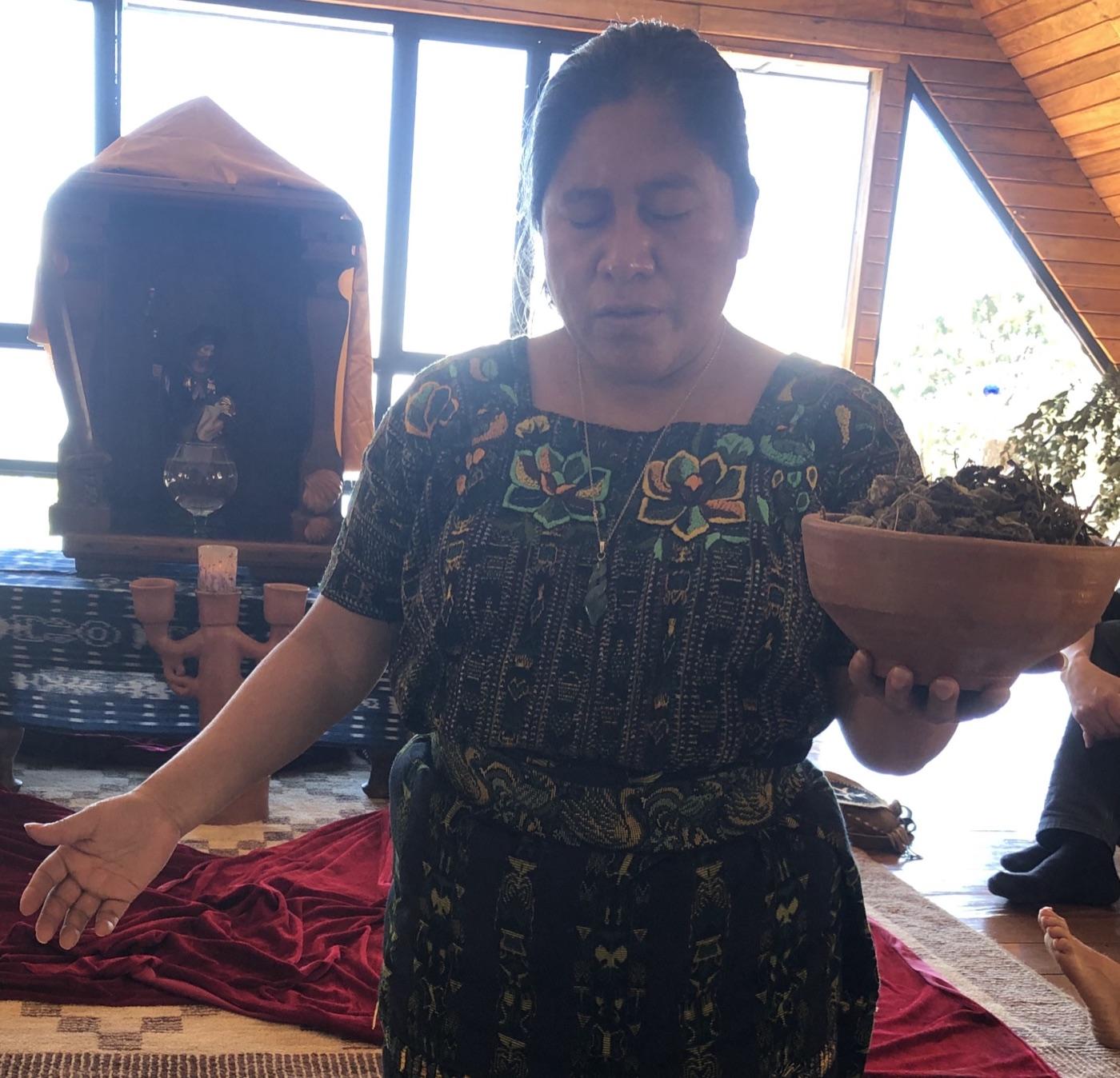 Carmen Vicente Cortéz in Maya-Tracht hält mit geschlossenen Augen eine Tonschale mit Heilkräutern, die andere Hand ausgebreitet, hinter ihr ein Altar